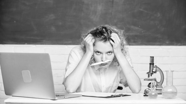 Black and white photo of a teach at her desk with her head on her hands, pencil in her mouth and a laptop on the desk. (Photo: Shutterstock)