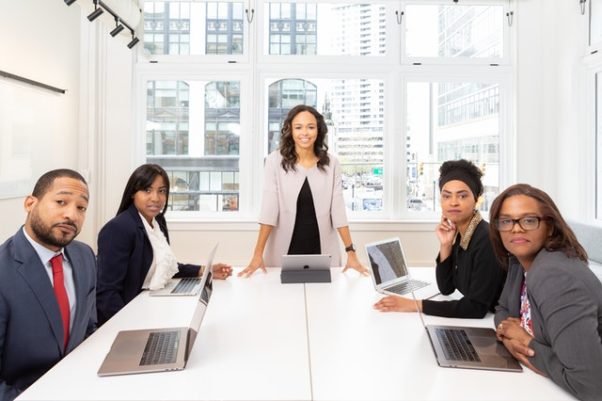 Woman standing in the center in a blazer with four people sitting around the table with laptops. (Photo: Rebrand City/Pexels)