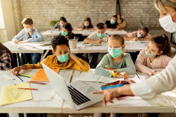 Students in a classroom wearing masks looking at a laptop with a masked teacher pointing to the screen. (Photo: Adobe Stock)
