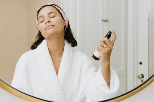 Woman in bathroom wearing a robe spraying moisturizer on her face. (Photo; Kalos Skincare/Upsplash)