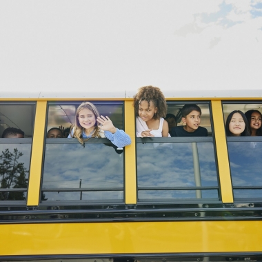 Medium wide shot of smiling middle school kids looking out open windows of school bus. (Photo: Penn State Health)