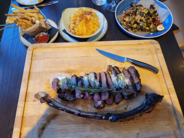 The three-course tomahawk ribeye steak dinner on a wooden cutting board in front with parmesan garlic fries (l to r), mac and cheese and crispy 
Brussels sprouts in white bowls behind. (Photo: Mark Heckathorn/DC on Heels)