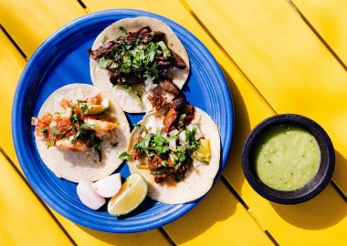 Three soft tacos and garnishes on a blue plate sitting on a yellow table with a cup of green sauce beside it. (Photo: Taqueria Habanero/Instagram)