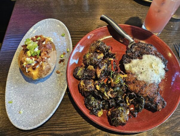 Ribeye with garlic-herb butter and Brussels sprouts on a red plate to the left and a loaded baked potato on an oval white plate on the right. (Photo: Nick Bullock/DC on Heels)