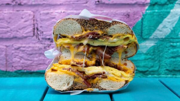 Two halves of a Call Your Mother's Sun 
City bagel sandwich piled on top of each other with bacon, spicy honey, an egg, American and cheddar cheeses on an everything bagel sitting in front of a pink wall on a teal table. (Photo: Call Your Mother/Facebook)