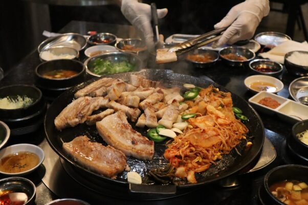 A waitress wearing gloves cooks meats, bean sprouts, jalepenos and garlic on a grill in the center of a table. The grill is surrounded by different sides and sauces in bowls. (Photo: Honey Pig Korean BBQ/Facebook)