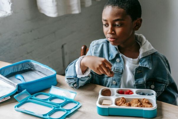 A young Black boy in a jean jacket and white T-shirt eats lunch out of a blue and white Bento-style lunch box. (Photo: Katerina Holmes/Pexels)