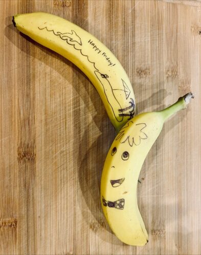 two bananas laying on a counter. One is decorated with a picture of a dolphin jumping out of water and an island with an umbrella and two palm trees that says "Happy Friday" and the other has hair, face and a bowtie drawn on it. (Photo: Dr. Kate Sanchez/Penn State Health)