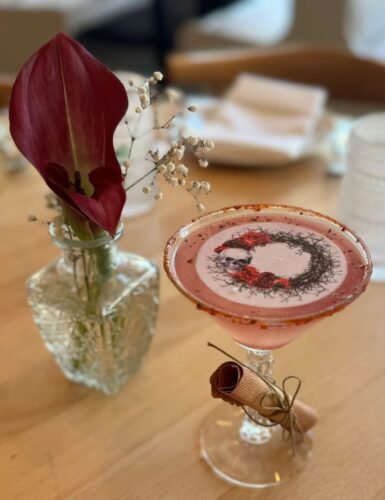 Ambar's blood moon margarita in a clear margarita glass with a bullseye pattern on top and a rolled note tied to the stem beside a deep red flower. (Photo: Ambar)
