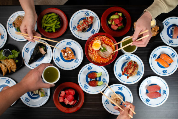 People eat sushi with chopsticks off blue and white plates and red bowls sitting on a wood table. (Photo: Emily J. Davis/Kura Sushi)