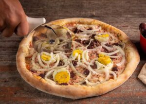A pizza topped with tuna, sliced onion, hard boiled egg, sauce and cheese is cut. (Photo: A Cantina Brasileira)