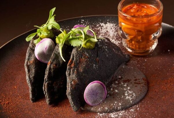 dLeña's empanada de langosta on a dark plate with microgreens and radish slices beside a drink in a skull glass. (Photo: dLeña)