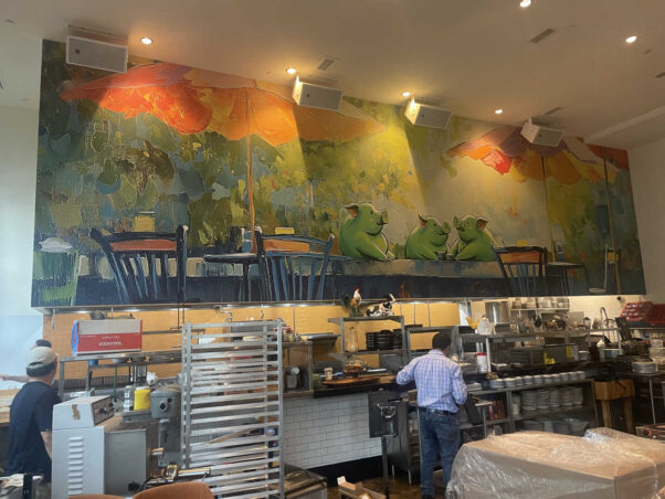 A mural above the open kitchen of three green pigs sitting at an outdoor bistro table, which two workers are in the kitchen below. (Photo: Katie taranto)
