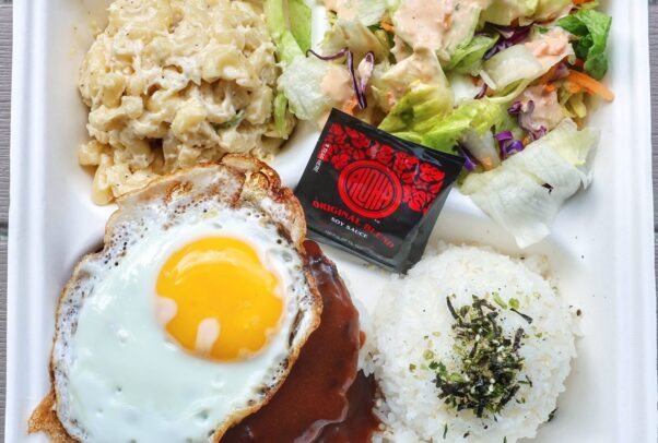 A loco moco lunch plate from Kau Kau with a hamburger steak topped with an sunnyside up egg in the lower left, a scoop of white rice in the lower right, a scoop of macaroni salad on the top left and a green salad in the top right with a black and red packet of soy sauce in the middle. (Photo: Kau Kau/Instagram)
