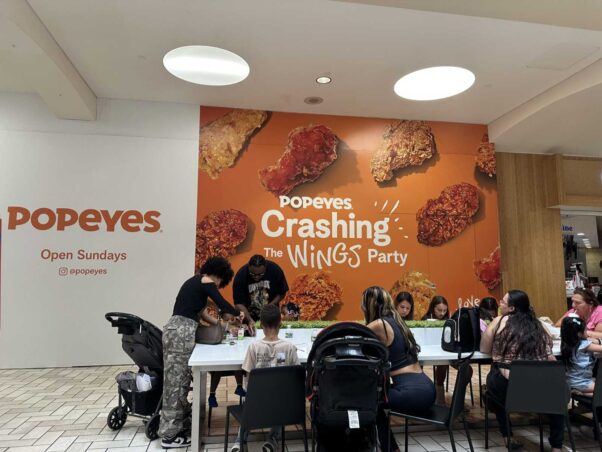 The future space of Popeye's at Tysons Corner Center's food court with people dining in front of a sign with a picture of fried chicken that says "Popeye's Crashing the Wings Party" and "Popeye's Open Sunday." (Photo: Angela
Woolsey)