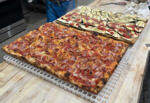 Two large square pies sit on cooling racks. One is covered in extra pepperoni and a white pizza with zucchini and anchovies. (Photo: Mustafa Durran)