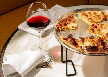 Osteria Mozza's focaccia di Recco — a bubbly, cheesy, cracker-like bread on a round pizza tray sitting on a wire rack on top of a white table cloth with a glass of red wine sitting beside it. (Photo: Birch Thomas)