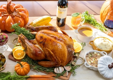 A whole roast turkey on a carving board with sage and lemon slices surrounded by by side dishes including gravy, cranberry sauce, stuffing and Brussels sprouts on a table covered with a white tablecloth, pumpkins and gourds. (Photo: Unconventional Diner)