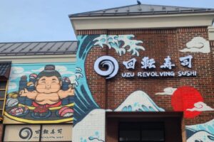 Exterior of Uzu Revolving Sushi in Rockville with a mural of a sumo wrestler in front of a conveyor belt with sushi, mountains and waves. (Photo: Amy Orndorff)
