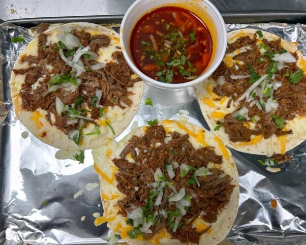 Three birria beef soft tacos with cheese, cilantro and onions on aluminum foil beside a cup of au jus. (Photo: Pho and Banh Mi Eatery Cafe/Instagram)