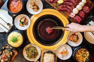 A hand uses tongs to cook a piece of beef on an in-table grill surrounded by various sides and a cutting board filled with beef, mushrooms and pineapple. (Photo: Seoul Prime)